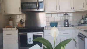 A kitchen with white cabinets, a stainless steel microwave, a stove, and a blender. There is a plant with a white flower in the foreground. - Corondo Shores Unique  & Beautiful Condo -  Stunning Views!!!  Pet Welcome! (Coronado)