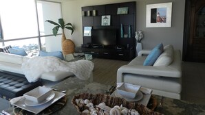 A modern living room with a large window, a flat-screen TV, and a comfortable seating area with a sofa and a couch. - Corondo Shores Unique  & Beautiful Condo -  Stunning Views!!!  Pet Welcome! (Coronado)