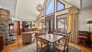 Un vaste salon doté d’une table à manger en bois, de sièges confortables, d’une cheminée en pierre et de grandes fenêtres offrant une vue sur l’extérieur.