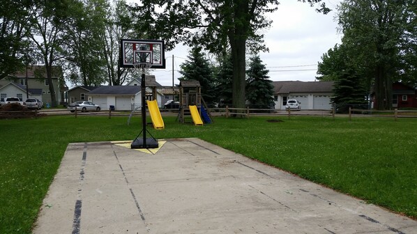 Een achtertuin met een basketbalveldje, twee glijbanen, een houten speeltuintje en een oprit van beton.