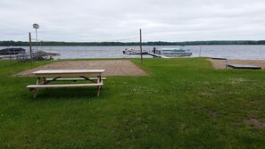 Ein Picknickplatz am Seeufer mit einer hölzernen Bank und einem kleinen Tisch, umgeben von grasbewachsenen Ufern und einem Steg mit Booten.