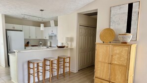 Una cocina moderna con armarios blancos, una isla central con taburetes, un mueble de madera con objetos decorativos y un cuadro abstracto grande en la pared.