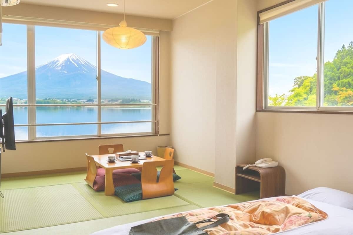 Una habitación con esteras de tatami, una cama, una mesa con sillas, una lámpara y vistas a la montaña a través de amplias ventanas.