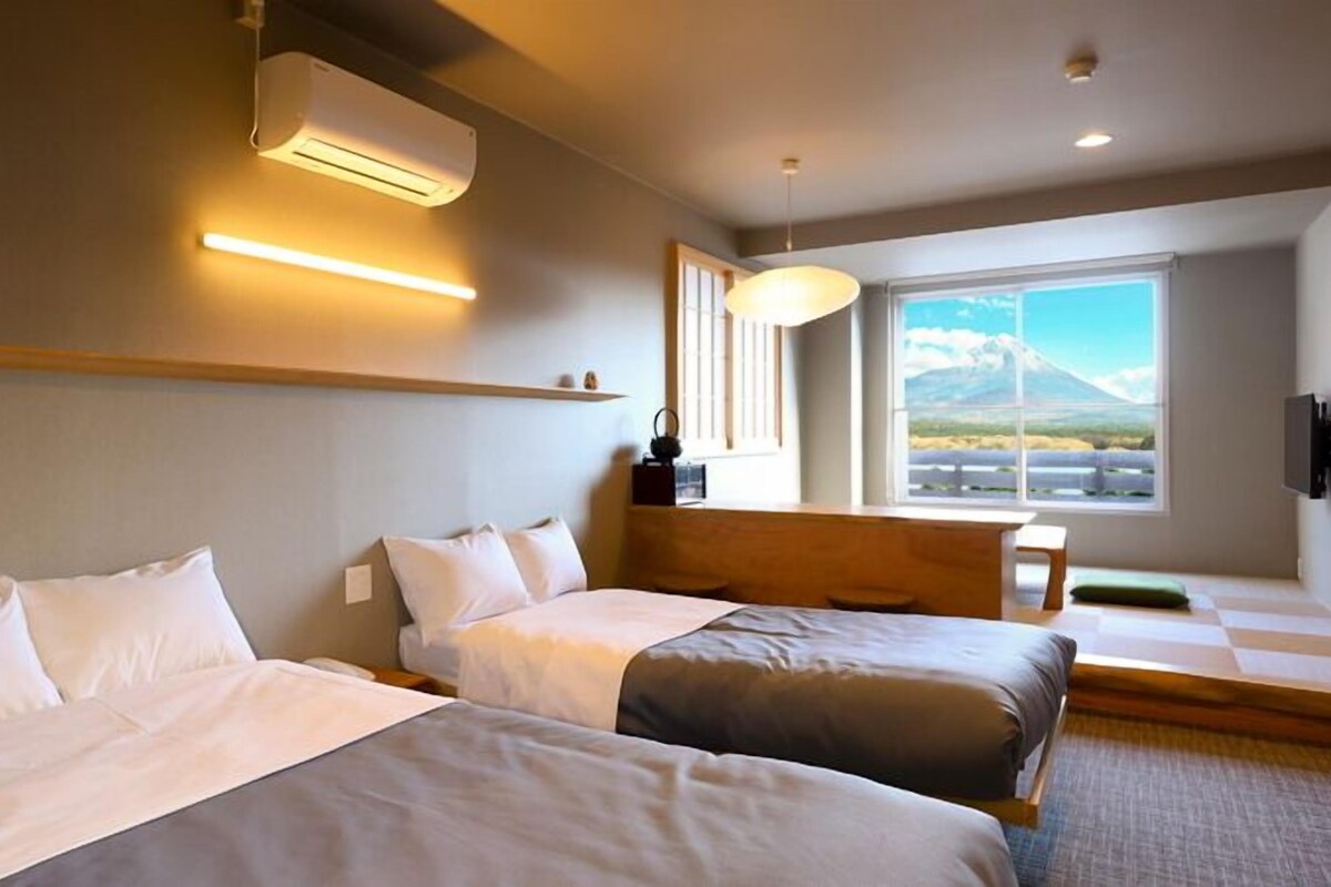Quarto de hotel com duas camas, uma janela grande com vista para as montanhas, ar condicionado e uma mesa com abajur.