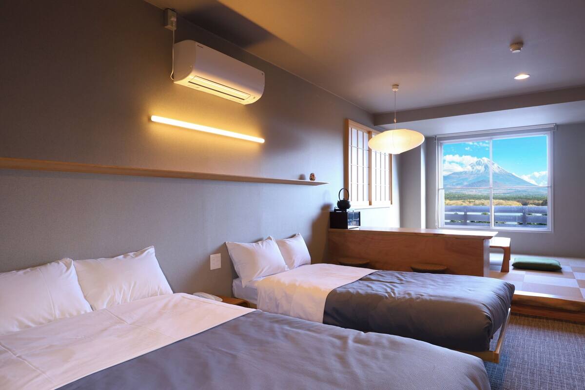 Habitación de hotel con dos camas, una ventana que ofrece vistas a la montaña, un aire acondicionado y una mesita de noche con lámpara.