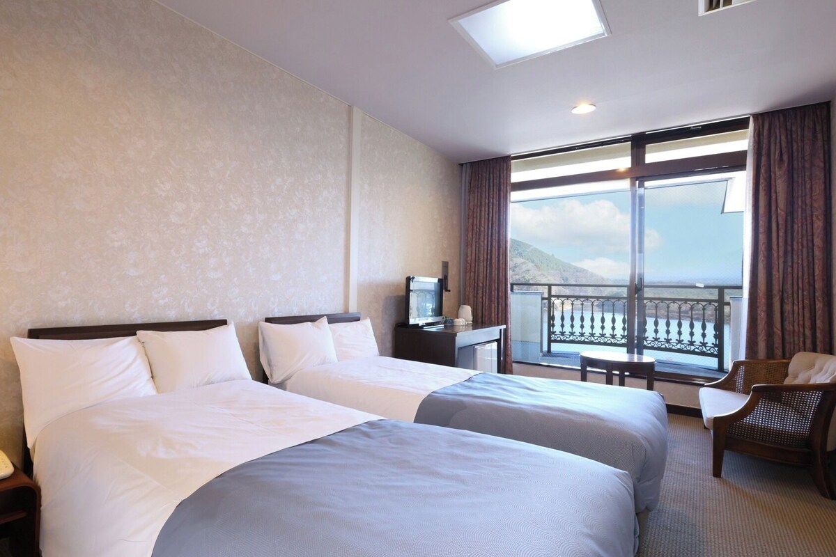 Quarto de hotel com duas camas, uma escrivaninha, uma cadeira, televisão e varanda com vista.