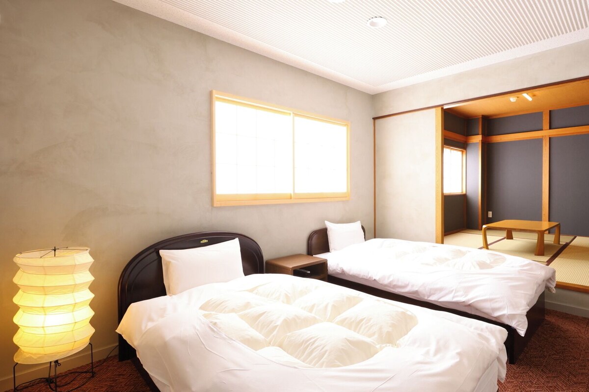 Quarto de hotel com duas camas, uma mesinha de cabeceira, um abajur e uma janela grande.