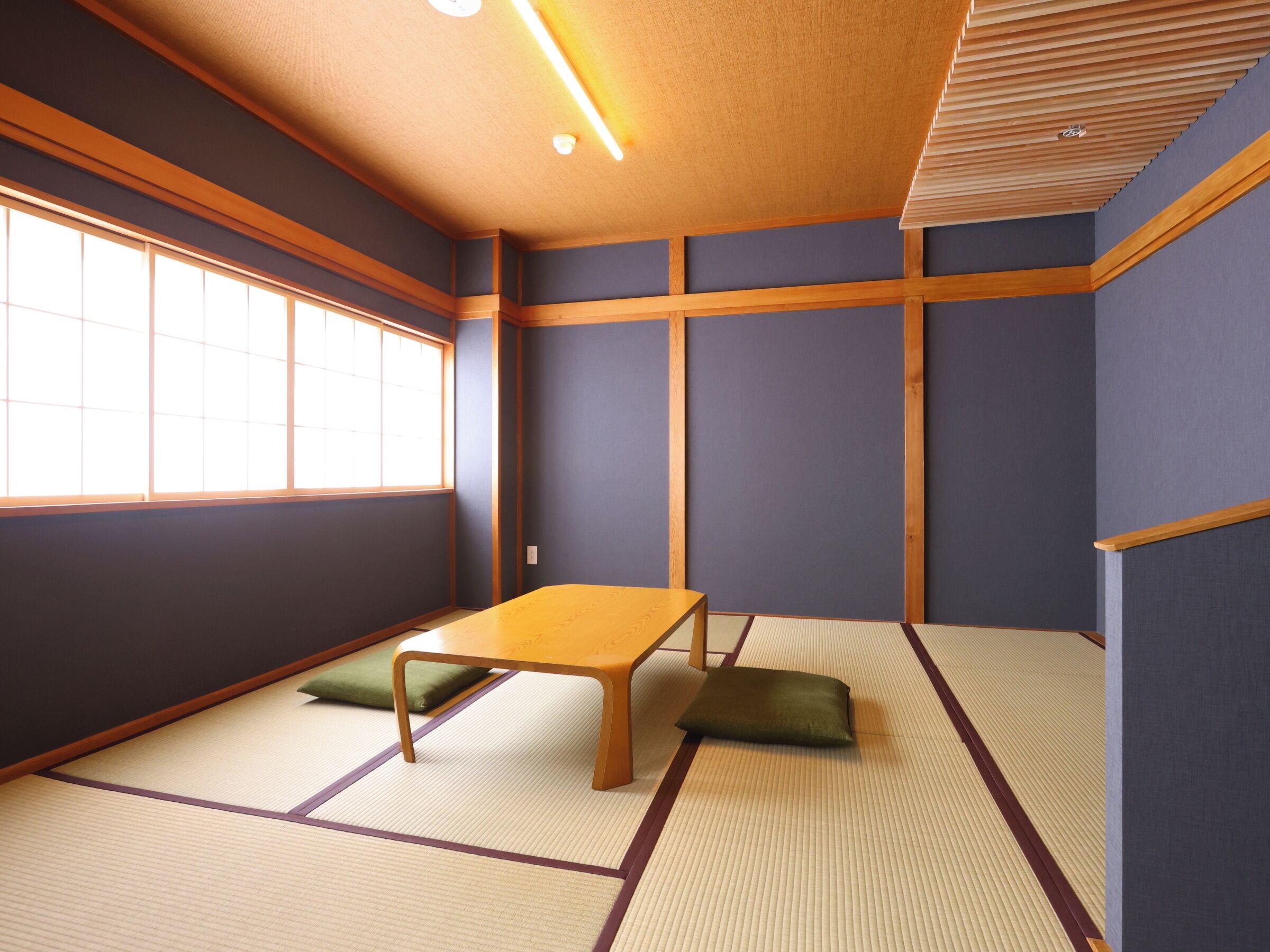 A tatami mat room with a low table and cushions, wooden beams on the walls, and a large window.