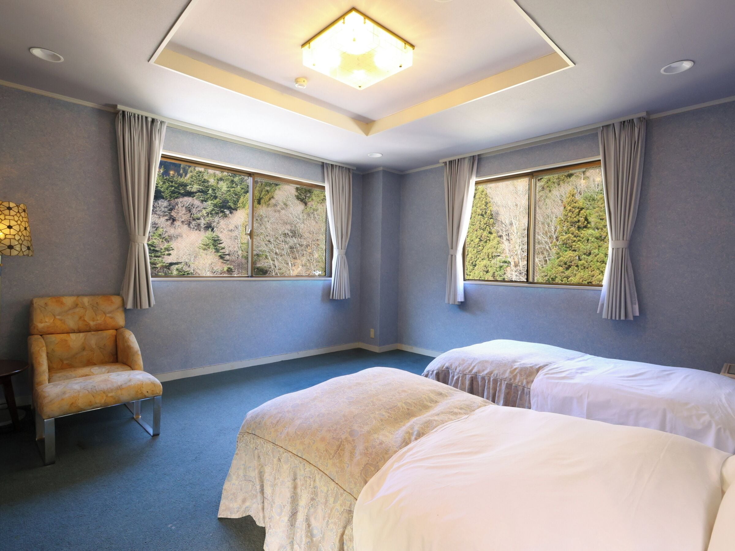 Una habitación con dos camas, una silla y ventanas amplias con cortinas.