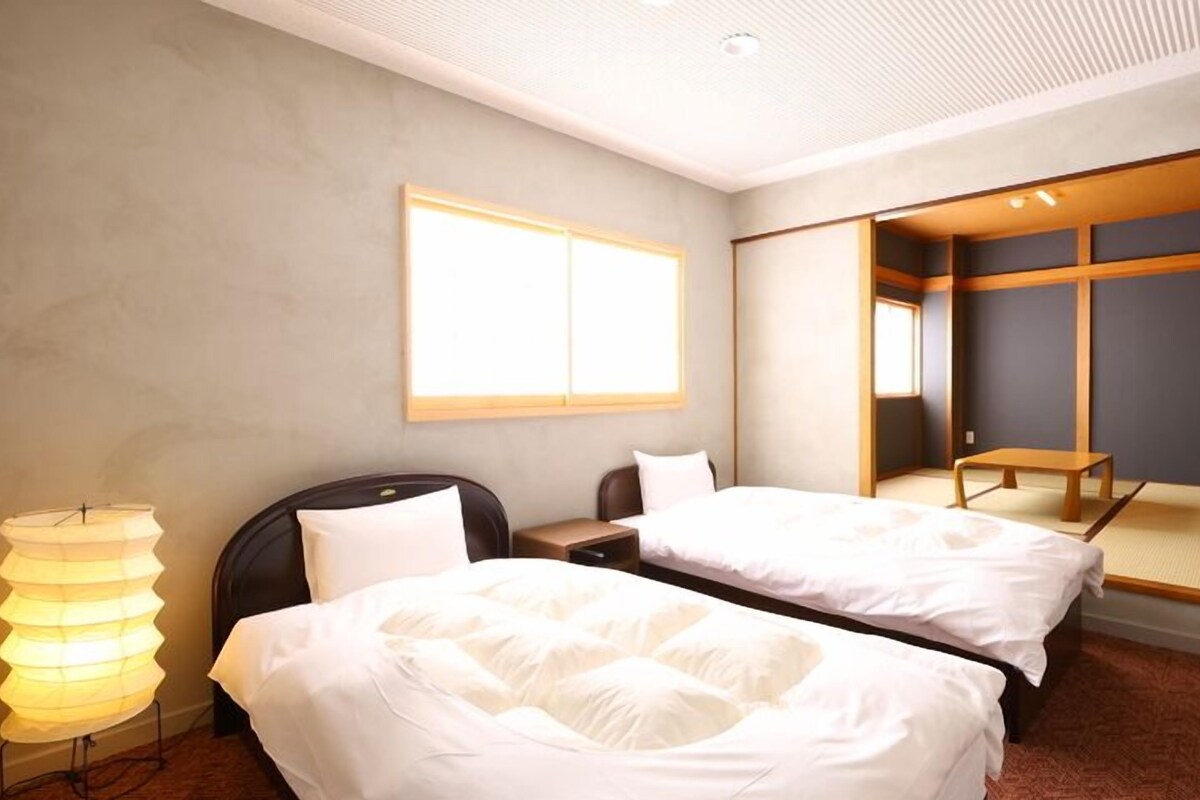 Quarto de hotel com duas camas, uma mesa de cabeceira, uma luminária e uma janela grande.