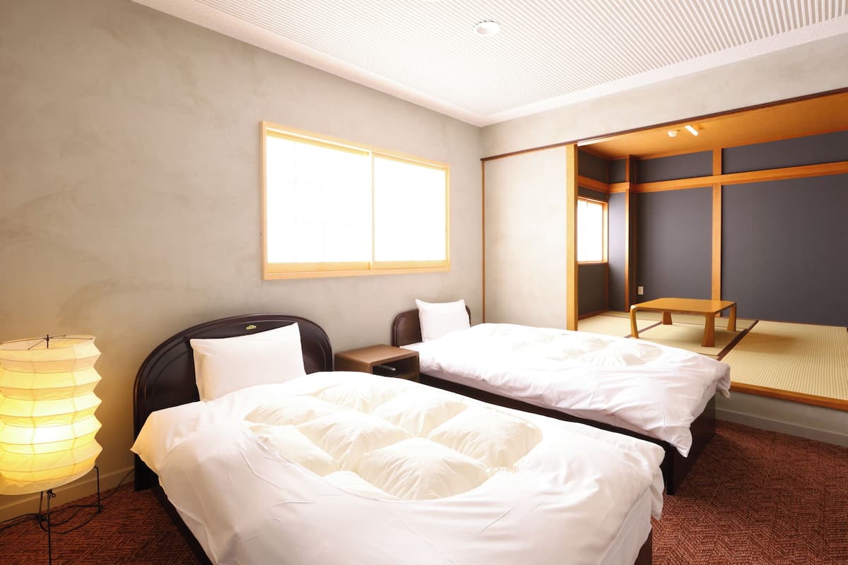 Habitación de hotel con dos camas, una mesita de noche, una lámpara, un espejo grande y vistas a una habitación tradicional japonesa.