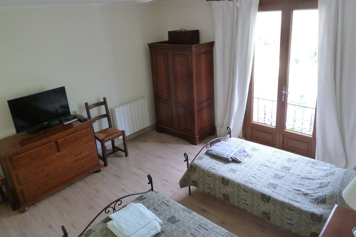 A bedroom with a wooden dresser, a television, a bed with a patterned bedspread, a chair, and a wardrobe.
