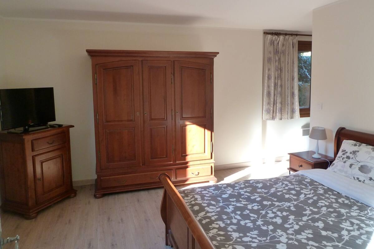 A bedroom with a wooden wardrobe, a bed with a floral bedspread, a nightstand with a lamp, and a window with curtains.