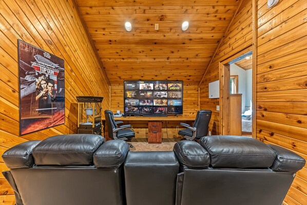 A wooden-paneled room with a flat-screen TV displaying various channels. There are two leather chairs in the foreground.