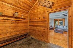 A wooden interior with a bench, hooks, and a door leading to a living area with a sofa and a ceiling fan.