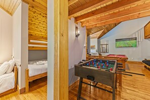 A cozy room with a wooden ceiling, a foosball table, a bed, and a television displaying a sports game. - 10 Min to Banner Elk | Sleeps 19 | Hot Tub, Sauna, Movie Theater & Mountain Views (Banner Elk)