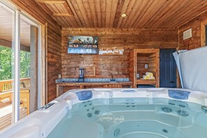 A wooden cabin interior with a hot tub, wooden walls, a mounted TV, and a door leading to another room.