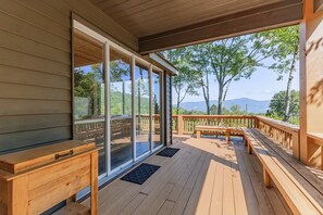 A wooden deck with a bench and a table, overlooking a forested area with a mountain in the distance.
