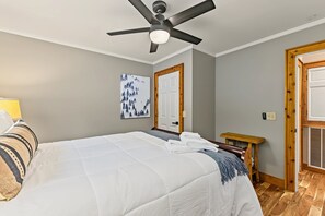 A hotel room with a bed, a ceiling fan, a painting, a door, and a small wooden bench.