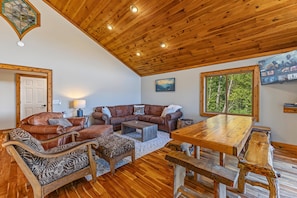 A cozy living room with a wooden ceiling, leather sofas, a wooden coffee table, and a large window with a view of trees. - 10 Min to Banner Elk | Sleeps 19 | Hot Tub, Sauna, Movie Theater & Mountain Views (Banner Elk)