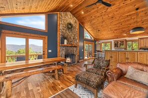 A spacious living area with a stone fireplace, wooden furniture, and large windows offering a mountain view. - 10 Min to Banner Elk | Sleeps 19 | Hot Tub, Sauna, Movie Theater & Mountain Views (Banner Elk)