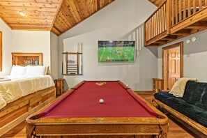 A room with a pool table, a bed, a sofa, and a television displaying a sports game.