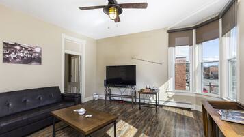 A living room with a black leather sofa, a wooden coffee table, a flat-screen TV on a stand, a ceiling fan, and large windows offering a view of the city.