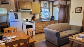 A compact kitchen with wooden cabinets, a stove, and a dining area with a table and chairs. - Cozy Fireside Cottage - The Rock (Nordegg)