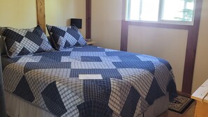 A bedroom with a bed, wooden beams, a ceiling fan, a window with curtains, and a small shelf. - Cozy Fireside Cottage - The Rock (Nordegg)