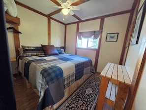 A bedroom with a bed, a ceiling fan, a window with curtains, and a wooden bench. - Cozy Fireside Cottage - The Rock King Cabin (Nordegg)