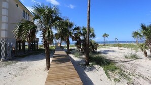 Ein hölzerner Weg führt zu einem Strand mit Palmen und einem Gebäude auf der linken Seite.