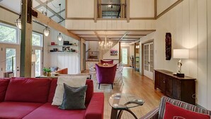A spacious loft-style living area with a high ceiling, wooden beams, a red sofa, a glass coffee table, and a purple armchair.