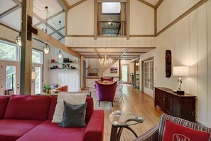 A spacious loft-style living area with a high ceiling, wooden beams, a red sofa, a glass coffee table, and a purple armchair.