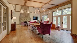 A spacious room with a wooden floor, a stone fireplace, a dining table with chairs, a sofa, and large windows.