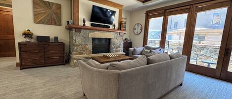 A living room with a stone fireplace, a flat-screen TV mounted above it, a large window with a view of the outside, and a wooden coffee table.