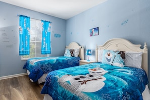 Un dormitorio con dos camas, cada una con ropa de cama azul a juego y un diseño de muñeco de nieve. Hay dos mesitas de noche con lámparas y una ventana con cortinas azules.