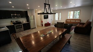 Una cocina moderna con armarios oscuros, electrodomésticos de acero inoxidable y una mesa de comedor de madera en un amplio salón con un ventanal y un sofá de cuero marrón.