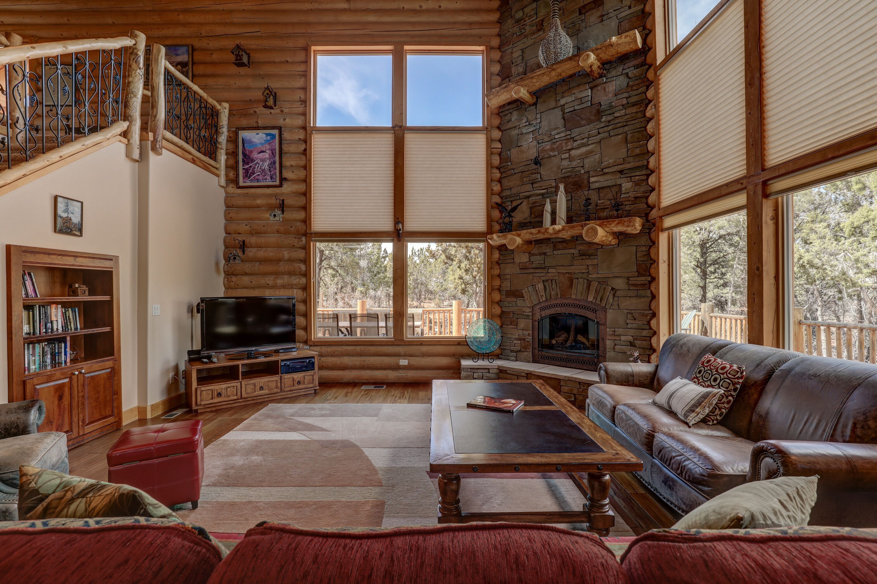 A log cabin interior with a stone fireplace, a large TV, a leather sofa, and a wooden staircase.