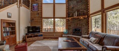 A log cabin interior with a stone fireplace, a large TV, a leather sofa, and a wooden staircase.