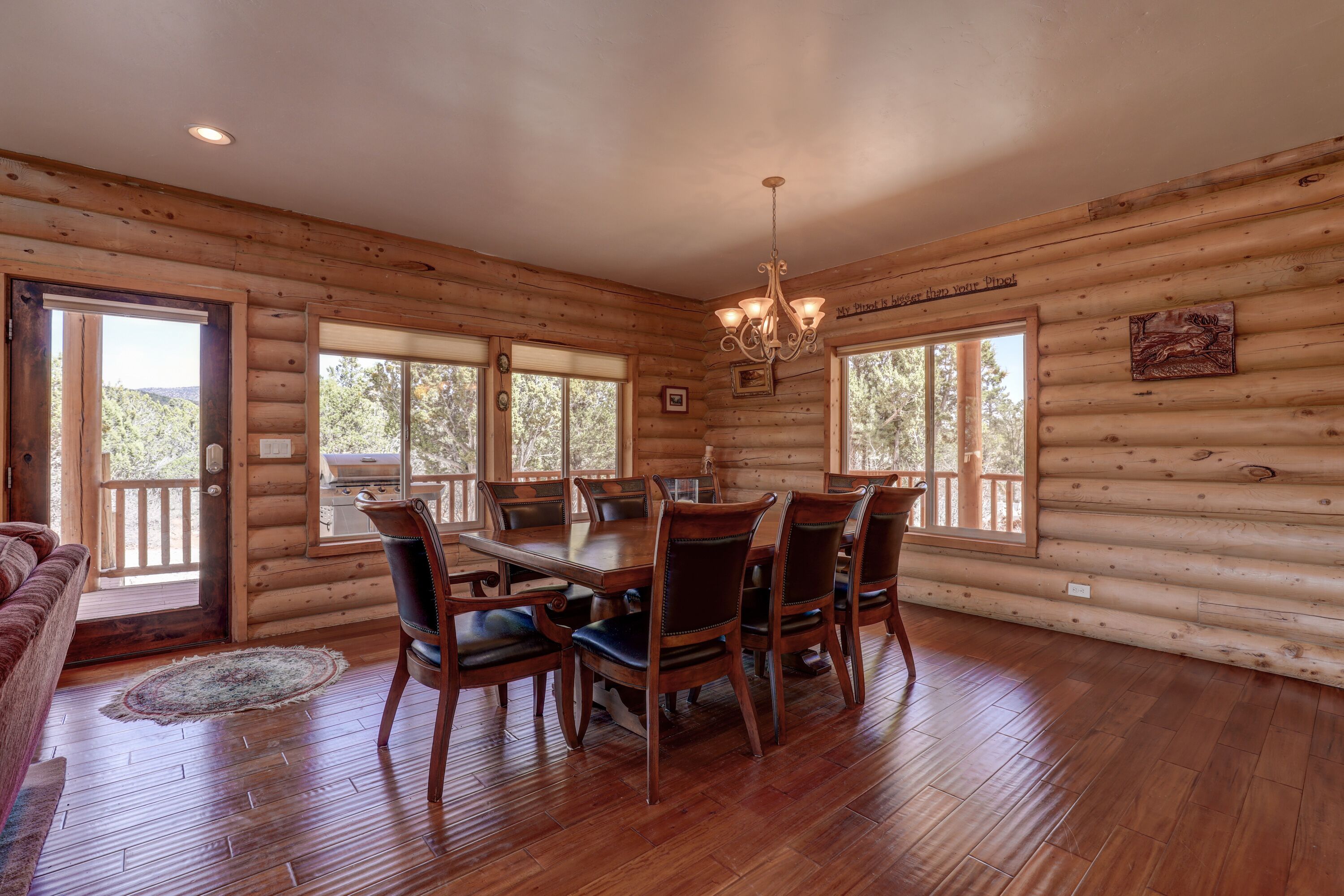 Interior de una cabaña de troncos con comedor, pisos de madera y ventanas grandes.