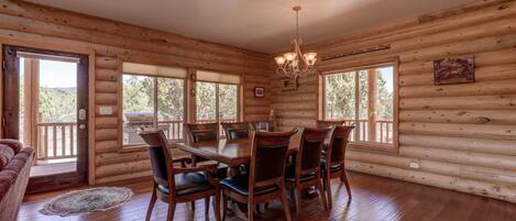 Interior de una cabaña de troncos con comedor, pisos de madera y ventanas grandes.