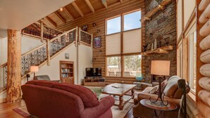 A log cabin interior with a wooden staircase, a red sofa, a wooden coffee table, and a stone fireplace.
