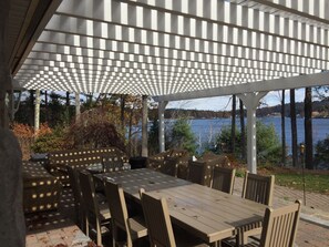 Un patio al aire libre con pérgola, mesas y sillas de madera, y vistas a un lago.