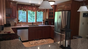 A kitchen with wooden cabinets, a stainless steel refrigerator, granite countertops, a window with a view of trees, and a hanging light fixture.
