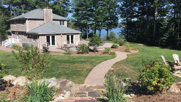 A stone house with a covered patio, a brick pathway, and a view of a lake.