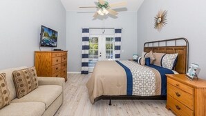 A bedroom with a bed, a wooden dresser, a wooden nightstand, a ceiling fan, a wall-mounted TV, and a door with curtains.