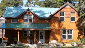 A two-story wooden cabin with a green metal roof, a front porch with outdoor seating, and a door with a small window. - 4 Bedroom Cottage On Manitoulin Island, Ontario (Providence Bay)