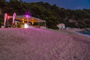A beachside restaurant with a covered outdoor seating area, overlooking the sea and a forested hillside.