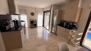 Cocina moderna con refrigerador, microondas y cafetera. Hay un fregadero, una encimera con tostadora y una pequeña mesa con una silla. La cocina tiene una puerta corrediza de vidrio que da a un patio.
