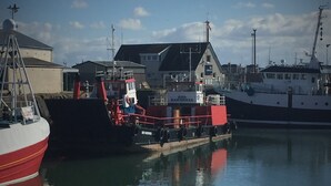 Un puerto con barcos amarrados, un edificio con el letrero 'RED BARONESS', y agua tranquila que refleja el cielo.